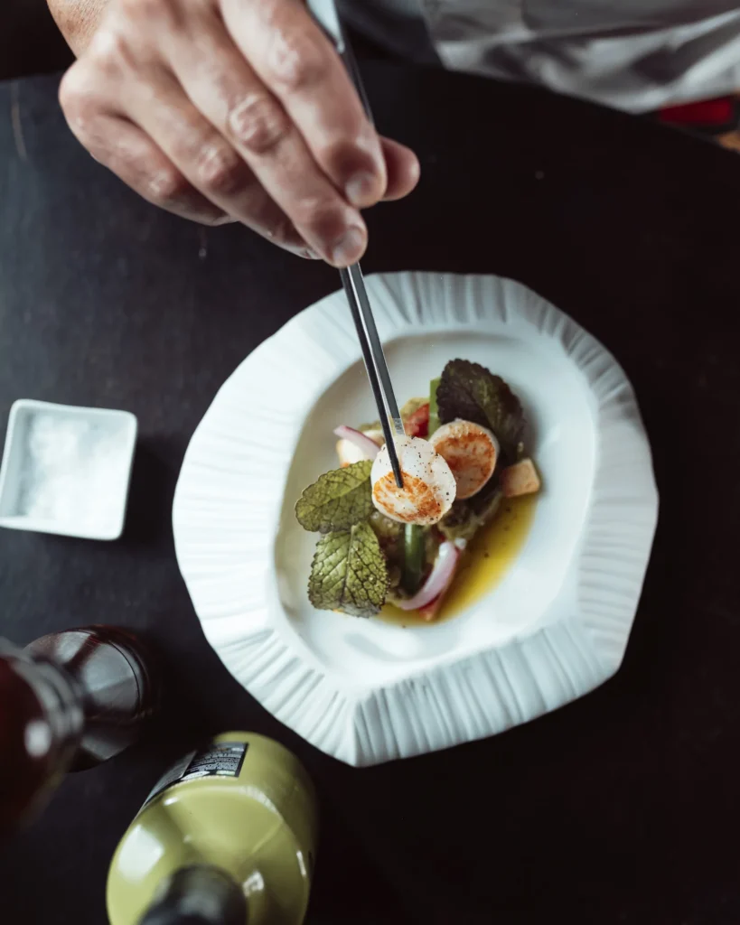 Manos del equipo terminan la presentación de un plato blanco con varios elementos.